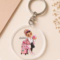 A clear acrylic keychain with a silver metal key ring, featuring a cartoon illustration of a girl with blonde hair, a pink suitcase, and the name 'Quinn' printed on it, displayed on a beige background with decorative dried flowers.