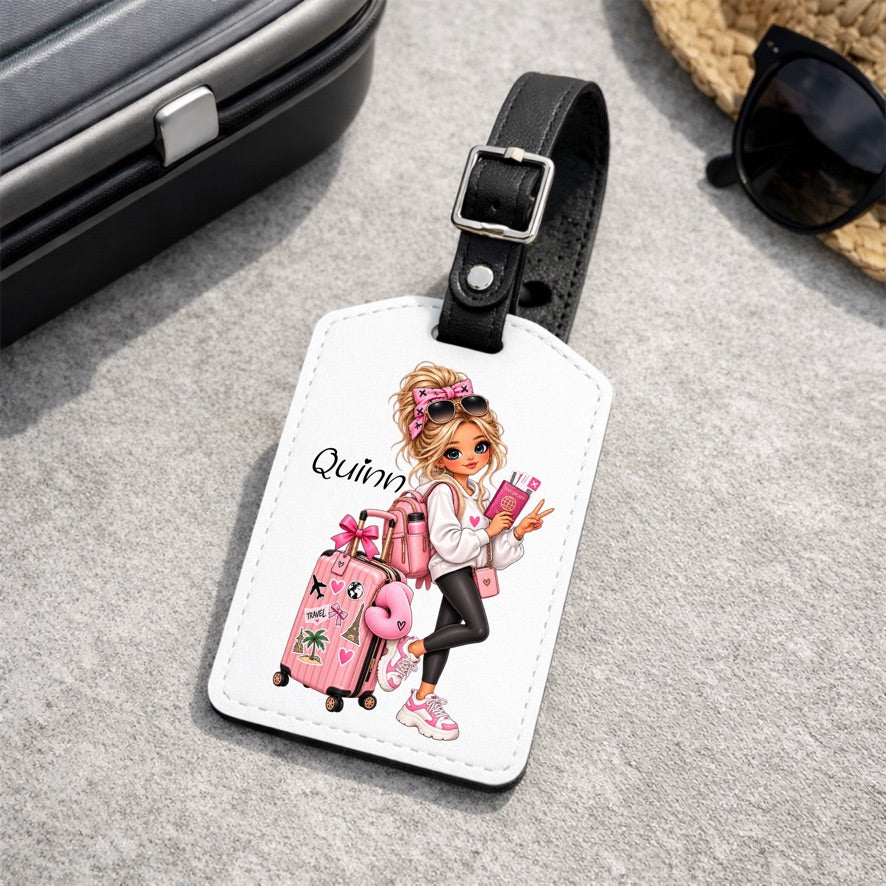 A white luggage tag with a black strap and silver buckle, featuring a cartoon illustration of a girl with blonde hair, pink accessories, and the name 'Quinn'. The tag is displayed on a gray textured surface with travel-themed props like a suitcase, sunglasses, and a straw hat.