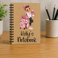 A personalized spiral-bound notebook with a kraft paper cover, featuring an illustration of a girl with a pink suitcase and the text 'Kelly's Notebook'. The notebook is displayed on a wooden desk with a blurred background containing a plant and a pencil cup.