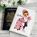 A personalized white faux-leather passport cover featuring a cartoon illustration of a blonde woman with a pink suitcase, pink backpack, and sunglasses, alongside the name 'Quinn'. It is styled next to a black British passport on a gray wood-textured background with white flowers.