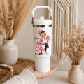 A white insulated travel tumbler with a lid and carry handle, held in a hand. The tumbler features a colorful illustration of a girl with pink luggage and the text 'Queen'.