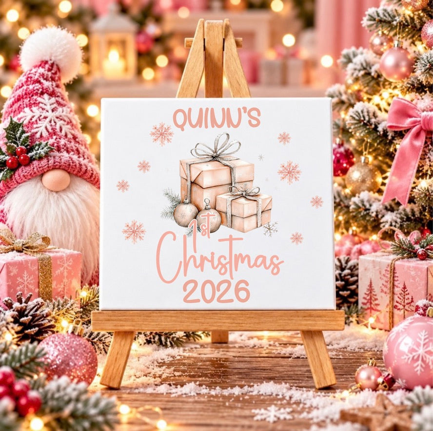 A square canvas print displayed on a small wooden easel, featuring a personalized Christmas design with the text 'QUINN'S 1st Christmas 2026' and an illustration of wrapped gifts. The scene is decorated for Christmas with a tree, ornaments, wrapped presents, and a gnome figure in the background.