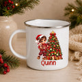 A white enamel-style mug with a silver rim, featuring a Christmas tree and a child in a Santa hat printed on the front, with the name 'Quinn' in red below. The mug is styled on a wooden surface with holiday greenery, red berries, and a burlap scarf in the background.