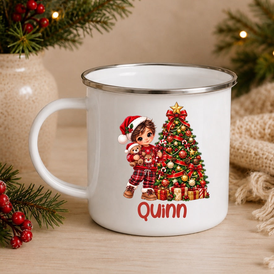 A white enamel-style mug with a silver rim, featuring a Christmas tree and a child in a Santa hat printed on the front, with the name 'Quinn' in red below. The mug is styled on a wooden surface with holiday greenery, red berries, and a burlap scarf in the background.