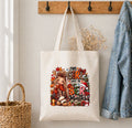 A cream canvas tote bag hanging on a wall hook, decorated with a colorful graphic of a woman in a plaid coat surrounded by autumn and holiday elements with the text 'THIS GIRL LOVES THE BE Months'. A denim jacket hangs on the right, a woven basket on the left, and dried flowers rest against the wall.