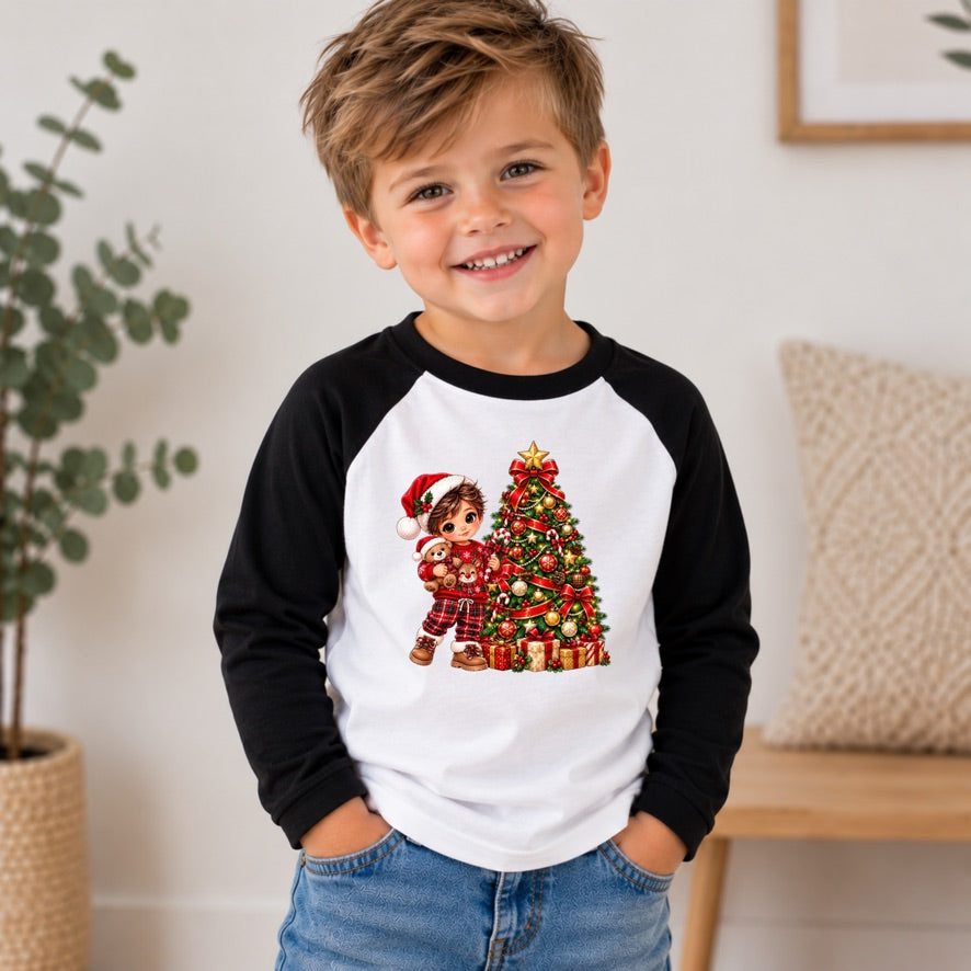 A smiling young boy wearing a black and white raglan baseball t-shirt with a Christmas graphic print of a boy and a decorated tree, paired with blue jeans in a bright indoor room.