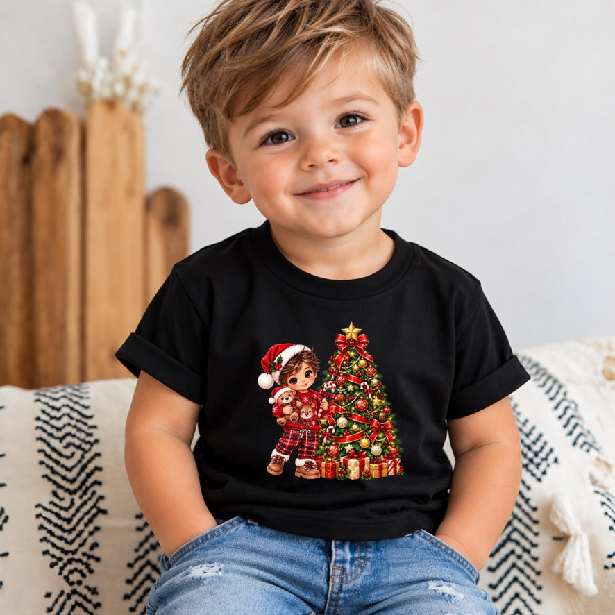 A smiling young boy wearing a black short-sleeve t-shirt with a festive Christmas graphic of a boy in a Santa hat hugging a decorated Christmas tree. He is sitting on a patterned throw blanket in a cozy indoor setting.