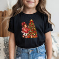 A child wearing a black short-sleeve t-shirt with a colorful Christmas-themed graphic print featuring a doll and a decorated Christmas tree, paired with blue jeans.