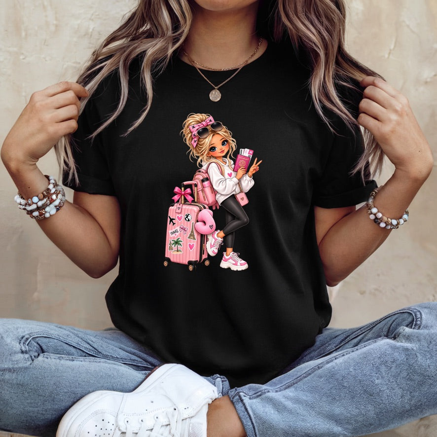 A woman wearing a black short-sleeve graphic t-shirt with a cartoon illustration of a girl with blonde hair, pink luggage, and a pink suitcase. She is sitting cross-legged in blue jeans and white sneakers against a textured beige wall.