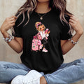A woman wearing a black short-sleeve graphic t-shirt with a cartoon illustration of a girl with blonde hair, pink luggage, and a pink suitcase. She is sitting cross-legged in blue jeans and white sneakers against a textured beige wall.