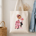 A cream canvas tote bag hanging on a wall, featuring a colorful cartoon illustration of a girl with pink luggage and accessories. The bag is styled in a home setting next to a woven basket, dried flowers, and a denim jacket.